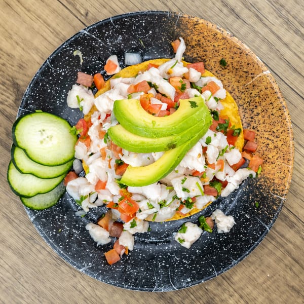 Ceviche tostada with avocado slices, diced tomatoes, and cucumbers on a speckled plate.