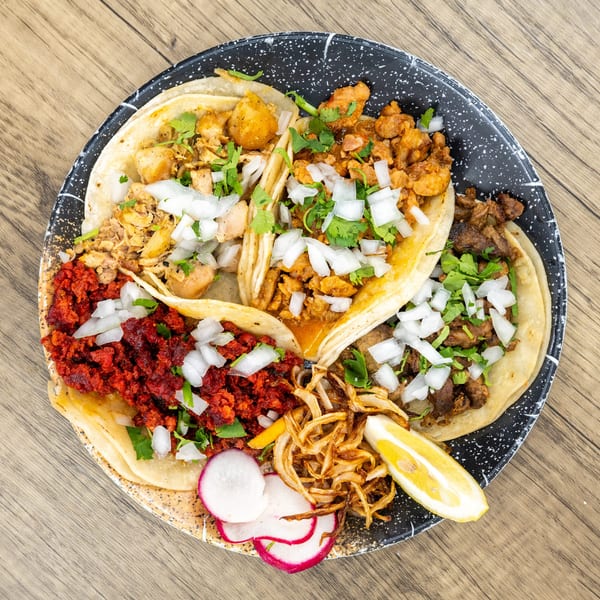 Four tacos with various fillings, onions, cilantro, fried onions, radishes, and a lemon wedge.