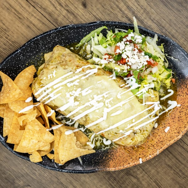 Two cheesy burritos covered in green sauce, with a side of tortilla chips and fresh salad.