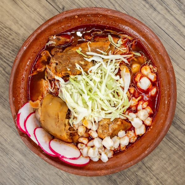 Overhead view of a pozole rojo in a clay bowl, garnished with cabbage, radishes, and hominy.